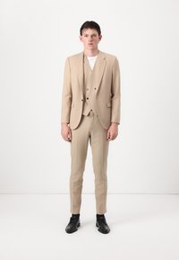 Abito beige su misura con giacca a un bottone, gilet coordinato e pantaloni slim-fit, abbinato a scarpe nere stringate.