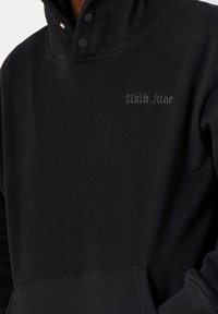 Schwarzer Hoodie mit Knopfkrawatte und "Sixth June" in kleinen grauen gotischen Buchstaben auf der Brust gestickt.