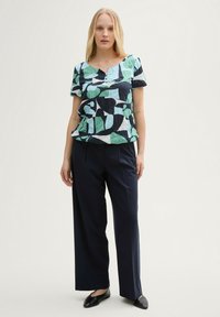T-shirt à manches courtes imprimé, avec un fond bleu marine orné de motifs de feuilles abstraites vertes et blanches. Accompagné de pantalon large foncé.