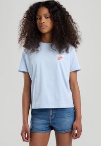 T-shirt azzurro chiaro a maniche corte con scollatura a girocollo e stampa grafica che recita "MONTAUK" in rosso. Indossato con pantaloni corti di jeans.