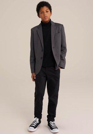 Grijze blazer, zwarte coltrui, zwarte broek en zwarte sneakers. De blazer heeft twee knopen en een voorzak; de casual stijl contrasteert met formele elementen.