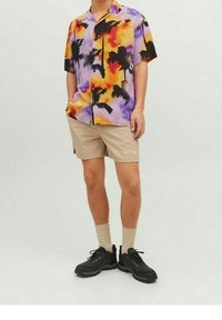 Camisa de manga corta con un estampado de palmeras multicolor en púrpura, naranja y negro; combinada con pantalones cortos beige y zapatos de senderismo negros.