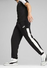 Schwarze Sportleggings mit weißen Seitenstreifen, einer figurschmeichelnden Silhouette und einem Logo am Oberschenkel, kombiniert mit grauen und weißen Turnschuhen.