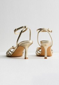 Sandali con tacco alto in metallo dorato, con cinturini curvi, chiusura con fibbia e superficie liscia. Presentano un tacco a stiletto sottile e un design aperto sulla punta.