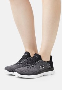 Scarpe da ginnastica nere e grigie con una tomaia texturizzata, lacci elastici e suola bianca. Presentano un logo sul lato e un design aerodinamico.
