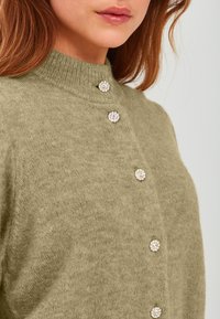 Olivno zelena pletena jopica z visokim ovratnikom, okrašena z okroglimi, prozornimi kamenčki, podobnimi gumbu, po sprednjem delu.