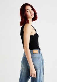 Melna bez piedurknēm blūze ar mežģīņu apdari, kas apvienota ar gaiši zilu džinsu biksēm ar ādas birku. Modelis pozē sānis, parādot apģērbu.