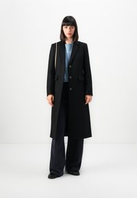 Donna con capelli scuri e lisci, che indossa un lungo cappotto nero con bottoni, una camicia blu, pantaloni larghi blu scuro, scarpe nere e una borsa a tracolla beige.