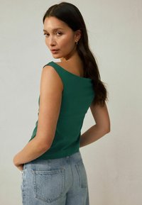 Mujer con cabello largo y oscuro, vistiendo una blusa sin mangas verde y jeans de cintura alta azul claro, posando con las manos en los bolsillos traseros.