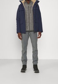 Veste à capuche marine avec doublure en fourrure beige, polaire zippée grise en dessous, pantalons gris clair et bottes de randonnée noires avec des accents de lacets.