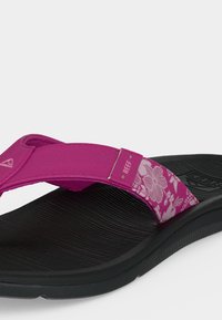 Μαύρη υφασμάτινη σόλα σαγιονάρων με φαρδιά ροζ λουράκι που έχει λευκά λουλουδάτα σχέδια και το λογότυπο της μάρκας Reef.