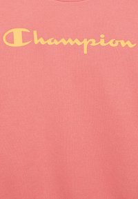 Felpa rosa realizzata in morbido tessuto con una texture a coste, caratterizzata da un grande logo giallo "Champion" in stile corsivo sulla parte anteriore.