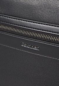 Détail d'un sac en simili cuir noir texturé avec une fermeture éclair argentée et un logo Calvin Klein en relief sur une bande lisse.