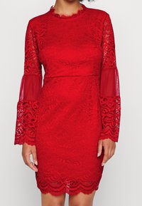 Röd spetsklänning med långa genomskinliga ärmar. Har scalloped kanter, blommönster och en figurnära design som slutar ovanför knäet.