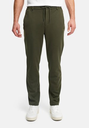 Man die olijfgroene joggerbroek met trekkoord in de taille en witte sneakers draagt, staand tegen een effen witte achtergrond.