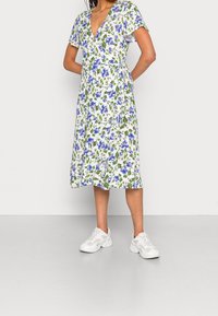 Robe portefeuille fleurie avec un fond blanc orné de fleurs bleues et vertes, manches courtes et décolleté en V profond, associée à des baskets blanches.