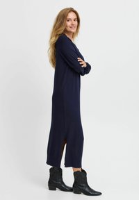 Tengerészeti hosszúujjú ruha oldalsó hasítékokkal és bordázott mandzsettával, fekete cowboy csizmával párosítva. Simaság és minimalista design jellemzi.