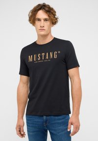 Czarna bawełniana koszulka z krótkim rękawem, okrągłym dekoltem i nadrukowanym złotym logo, w połączeniu z niebieskimi dżinsami.