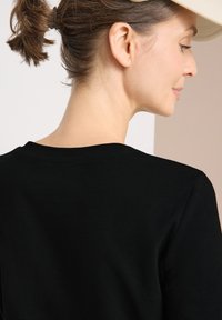 Schwarzes Baumwoll-T-Shirt mit rundem Halsausschnitt, kurzen Ärmeln und einer glatten Textur. Der Hintergrund zeigt ein hellbeiges und weißes geometrisches Design.
