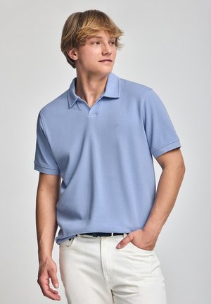 Polohemd aus hellblauem Stoff mit Kragen und kurzen Ärmeln, auf der Brust mit einem kleinen gestickten Logo. Getragen mit weißen Hosen.