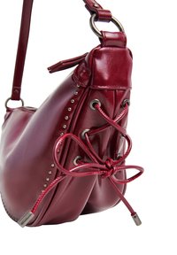 Κόκκινη δερμάτινη τσάντα ώμου με μεταλλικά καρφιά, λεπτομέρειες με κορδόνια στο πλάι, στρογγυλή μορφή και ρυθμιζόμενο λουρί με μεταλλικό δαχτυλίδι σε λευκό φόντο.