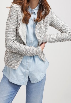 Cárdigan de punto gris jaspeado con un diseño de frente abierto, llevado sobre una camisa de botones azul claro, que presenta un corte relajado y una tela texturizada.