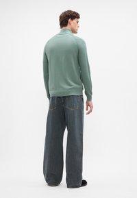 Minzgrüner Strickpullover mit hohem Kragen, kombiniert mit locker sitzenden dunkelblauen Jeans aus Denim, die über Rückentaschen verfügen.