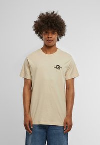 Beige cotton t-shirt with a round neck and short sleeves. Features black text logo on the chest. Loose fit and soft texture.