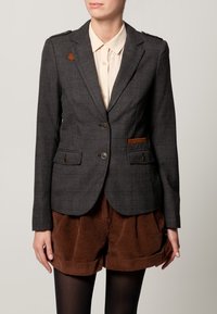 Femme portant un blazer à carreaux gris foncé avec des accents marron, une chemise beige à boutons, un short en velours côtelé marron et des collants noirs contre un fond neutre.