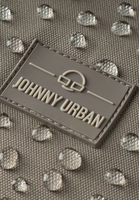 Szürke szövet, kiemelkedő vízcseppekkel és egy téglalap alakú logóval, amelyen a "JOHNNY URBAN" felirat neutrális árnyalatban van gravírozva. Texturált felület.