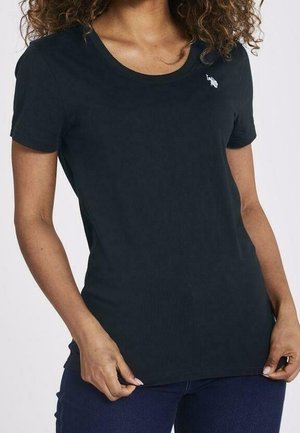 Femme portant un t-shirt noir uni à manches courtes avec un petit logo blanc sur la poitrine gauche, associé à un jean bleu foncé.