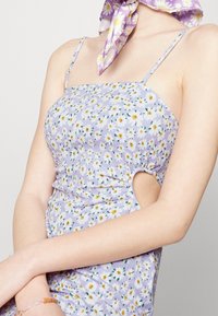 Robe florale lavande avec texture froncée, fines bretelles et un détail découpé sur le côté. Présente de petits motifs de fleurs jaunes et vertes.