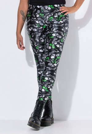 Person iført sorte leggings med mønster af hvide og grønne kranier og sorte kampstøvler, stående med den ene hånd på hoften.