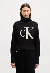 Maglione a collo alto lavorato a maglia nera con un grande logo bianco 'ck' al centro, con texture a costine e design crop.