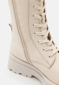 Krémové kožené kotníkové boty s robustní podešví, zdobené bočním zipem, šněrováním v přední části a kontrastním prošíváním.