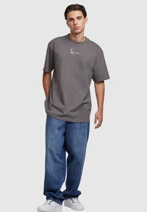 Graues Baumwoll-T-Shirt mit kurzen Ärmeln und Rundhalsausschnitt, versehen mit weißem Drucktext auf der Brust. Kombiniert mit lockeren blauen Jeans.
