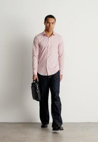 Helles rosa Hemd mit Knöpfen, dunkle Jeans, schwarze Schuhe, hält eine schwarze Lederakte, steht vor einem schlichten weißen Hintergrund.