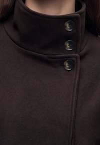 Gros plan sur un cou et une poitrine portant un manteau marron foncé avec un col montant fermé par trois grands boutons noirs.