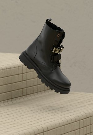 Stivale da combattimento in pelle nera con suola spessa, chiusura con lacci e due cinturini decorativi, sospeso sopra gradini in piastrelle beige.