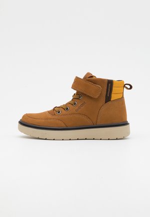 Bota infantil impermeable color marrón de caña alta con cordones en la parte delantera, correa de velcro, cuello acolchado amarillo y suela de goma beige.