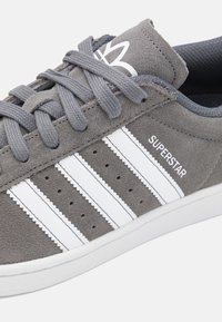 Šedé semišové tenisky s třemi bílými pruhy, perforovanými detaily a značením. Zaoblená špička a bílá gumová podrážka.