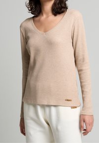 Camiseta de manga larga y escote en V en tejido acanalado beige con una textura sutil. Presenta un pequeño parche de logo cerca del dobladillo y se combina con unos pantalones blancos texturizados.