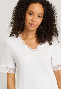 Valkoinen t-paita, jossa on V-pääntie sekä pitsireunus ja pitsikoristeet lyhyissä hihoissa. Silkkinen rakenne, rento muotoilu.