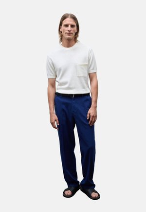 Jeune homme aux cheveux mi-longs portant une chemise blanche à manches courtes, un pantalon bleu, une ceinture noire et des sandales noires, debout devant un fond uni.