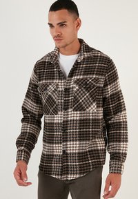 Cămașă în carouri maro și alb, confecționată dintr-un material textil, având două buzunare pe piept, închidere cu nasturi și un guler clasic.