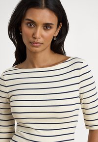 Žena s tmavými vlasmi, ktorá nosí zlaté náušnice a krémové tričko s vodorovnými námorníckymi modrými pruhmi a tromištvrtinovými rukávmi, na bielom pozadí.