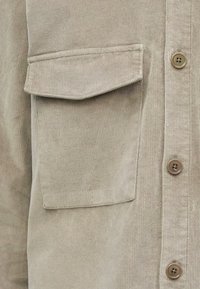 Beige corduroy overhemd met een borstzak, knoopsluiting aan de voorkant en vijf knopen. Getextureerde stof met verticale ribbels.