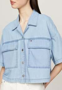 Light blue denim jacket with a cropped design, short sleeves, two chest pockets, and button-front closure. Features subtle stitching details.
