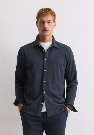 Marineblaue langärmlige Hemdbluse aus weichem Stoff mit spitzem Kragen und weißen Knöpfen, kombiniert mit einem weißen Unterhemd.