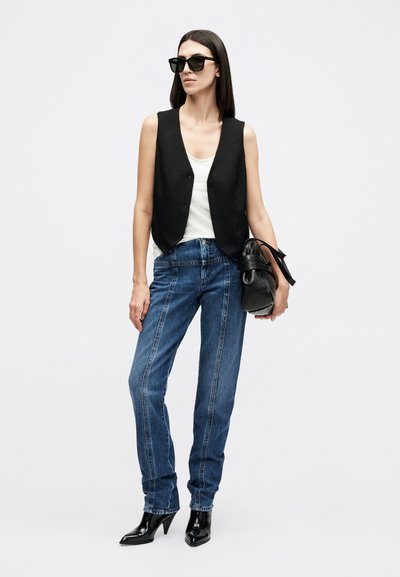 Γυναίκα με μαύρο αμάνικο γιλέκο, λευκό αμάνικο τοπ, μπλε τζιν, μαύρα μποτάκια με τακούνι, γυαλιά ηλίου, κρατώντας μια μαύρη τσάντα.